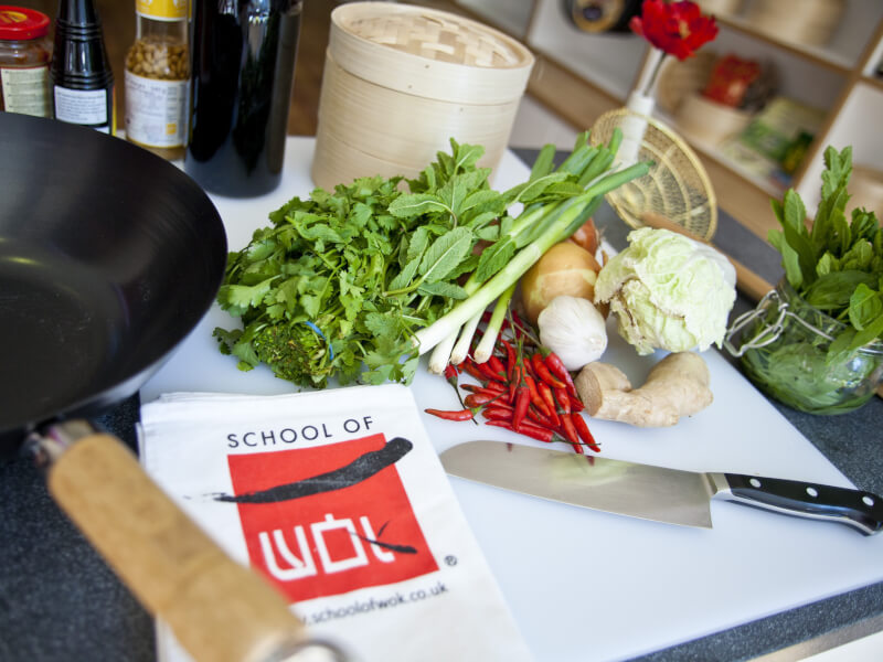 A large bunch of coriander and a selection of fresh vegetables sit by a wok, ready to cook
